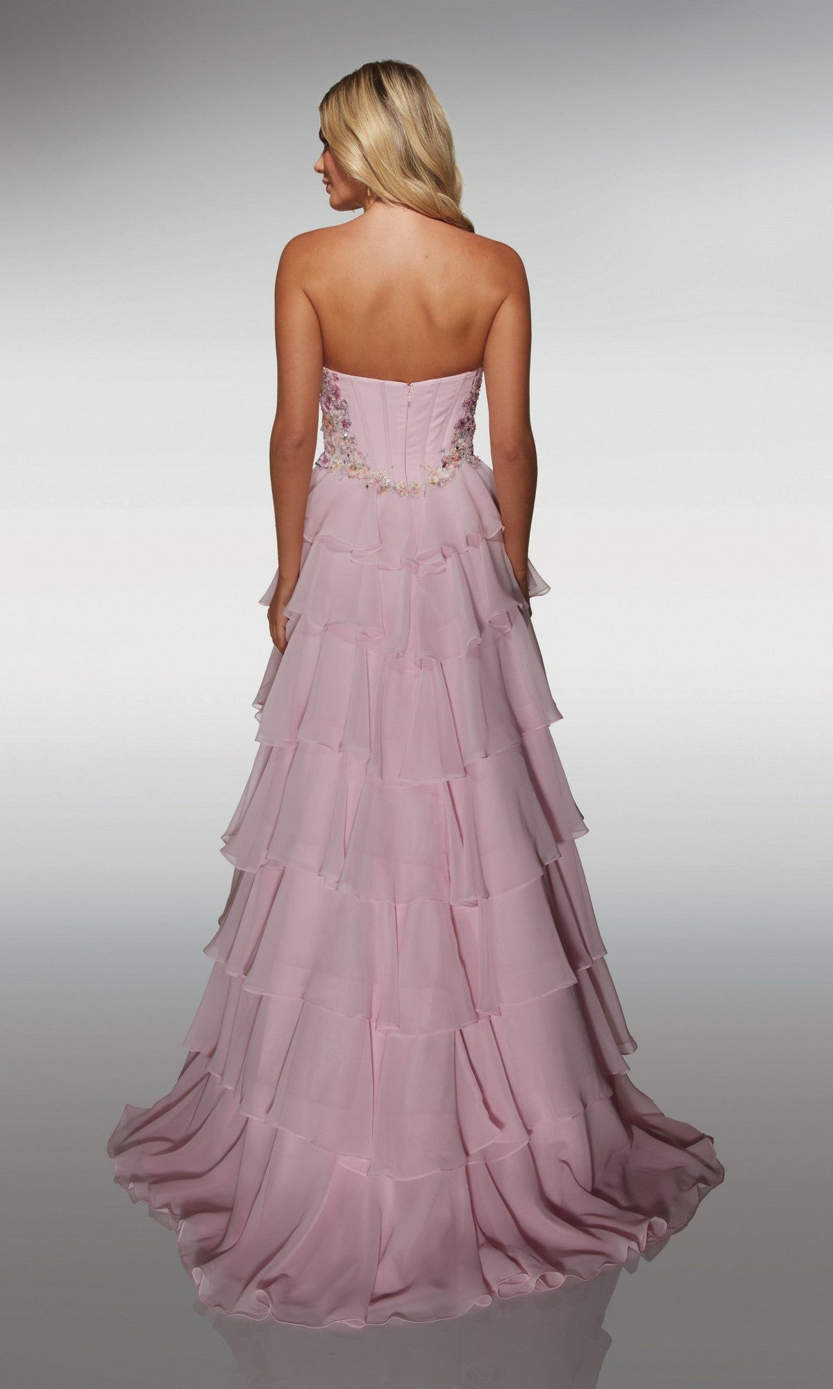 A woman with blonde hair, facing away, wears the Alyce 62013 long prom dress—strapless, floor-length chiffon with a tiered skirt and floral embroidery and appliques accenting the corset bodice.